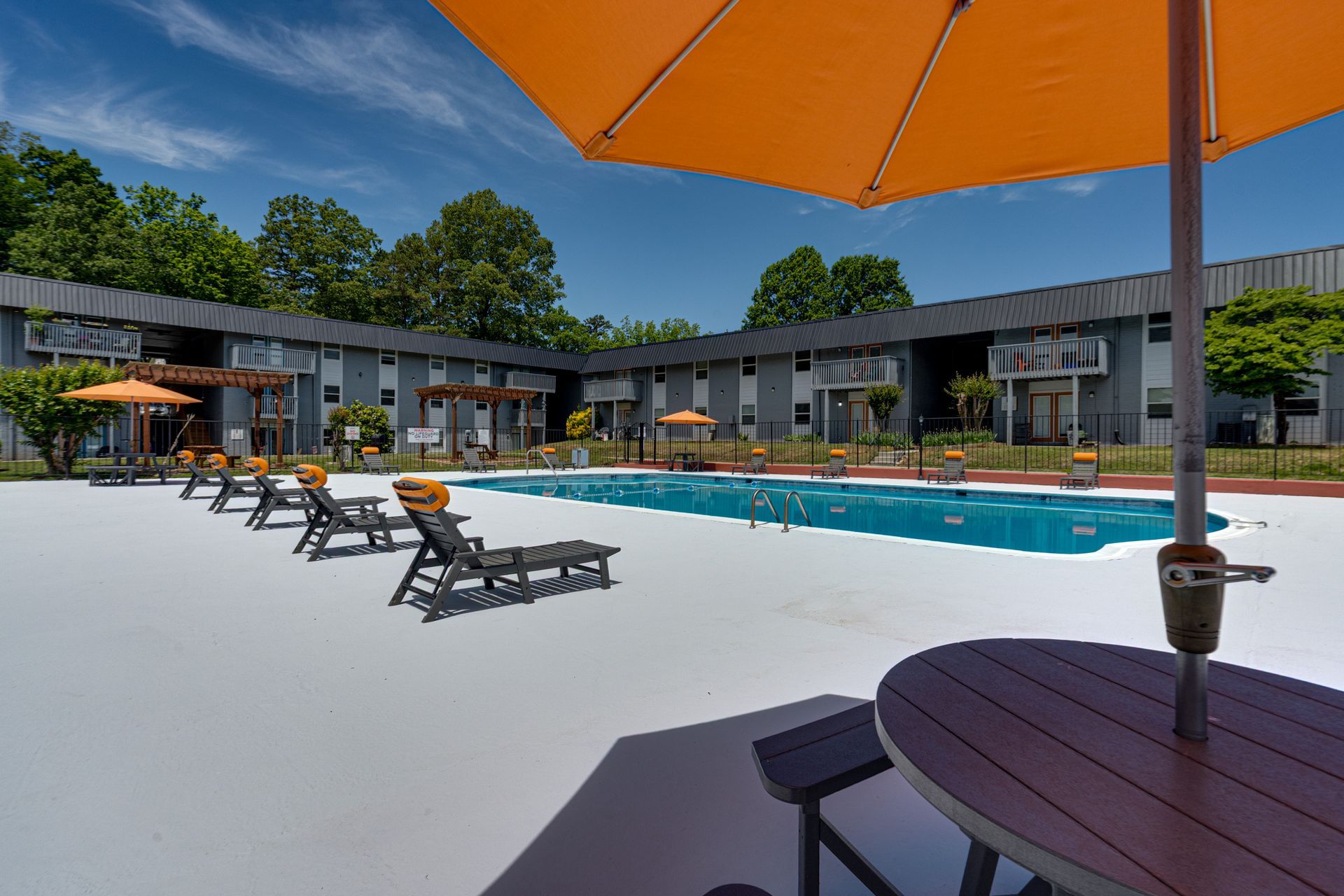 A bright blue swimming pool sits in front of a motel with lounge chairs, trees, and an orange umbrella under a sunny sky.