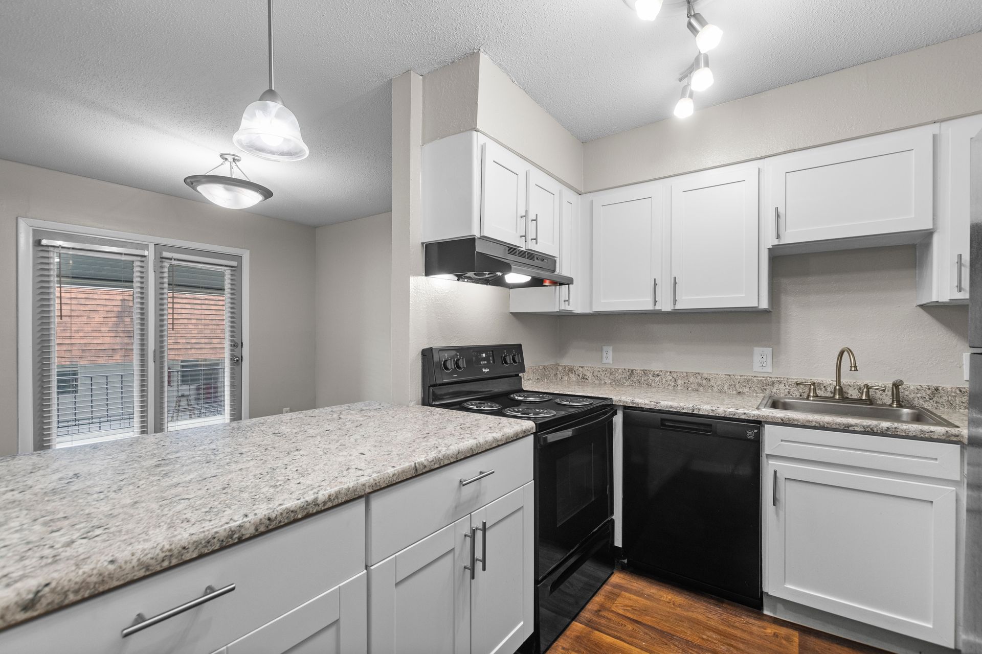 A kitchen with white cabinets, granite countertops, and black appliances, illuminated by pendant and track lighting.