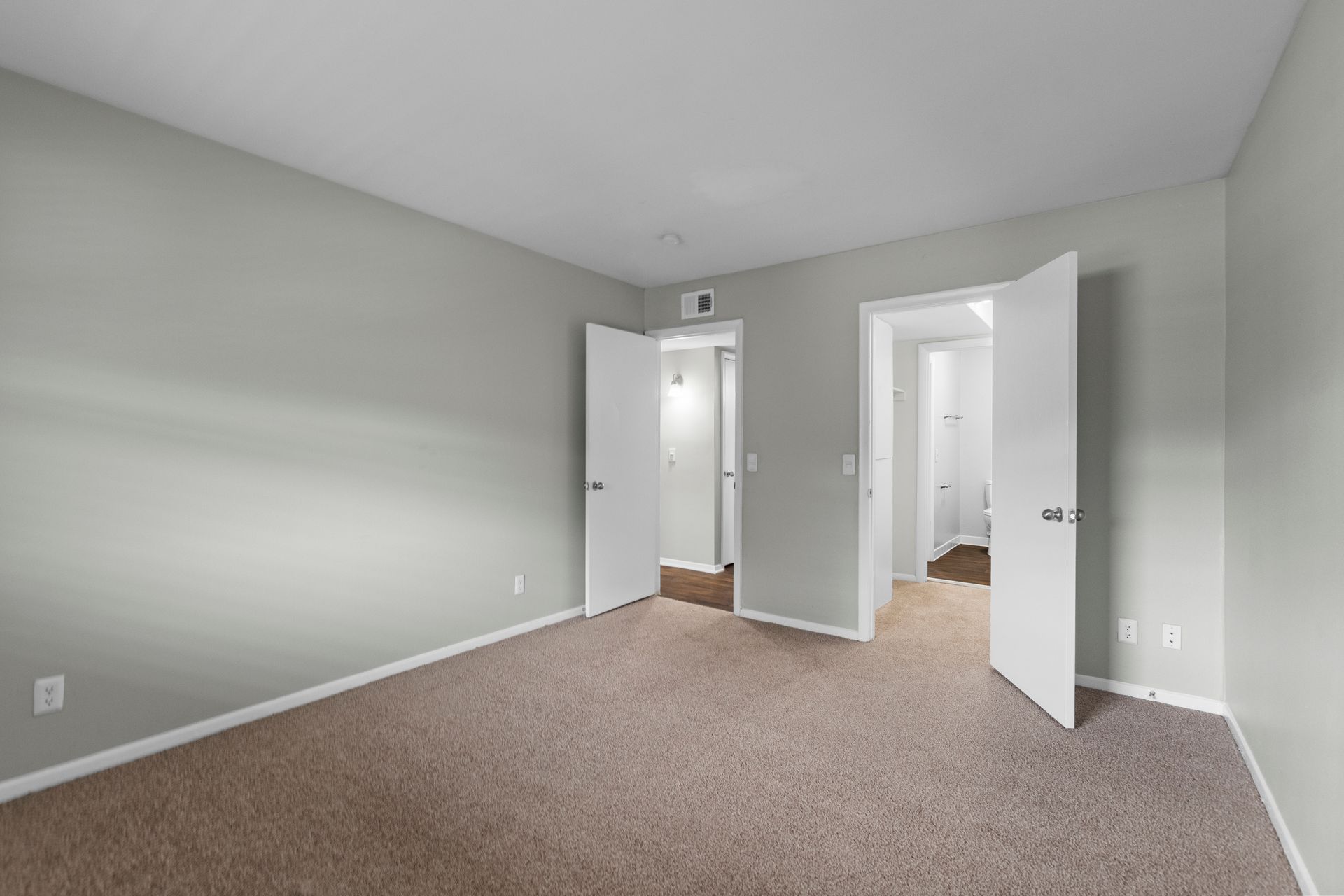 Empty room with light grey walls and beige carpet featuring two open doorways leading to other rooms.