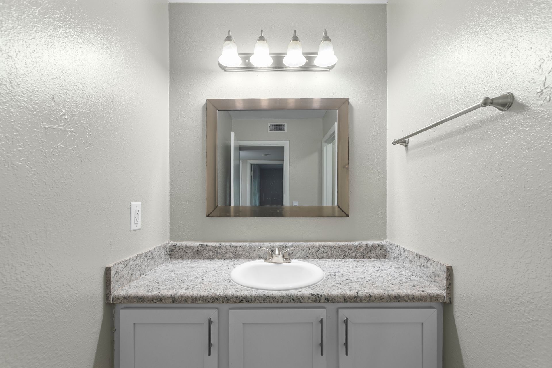 A bathroom vanity with a gray cabinet, granite countertop, white sink, rectangular mirror, and four-bulb light fixture.