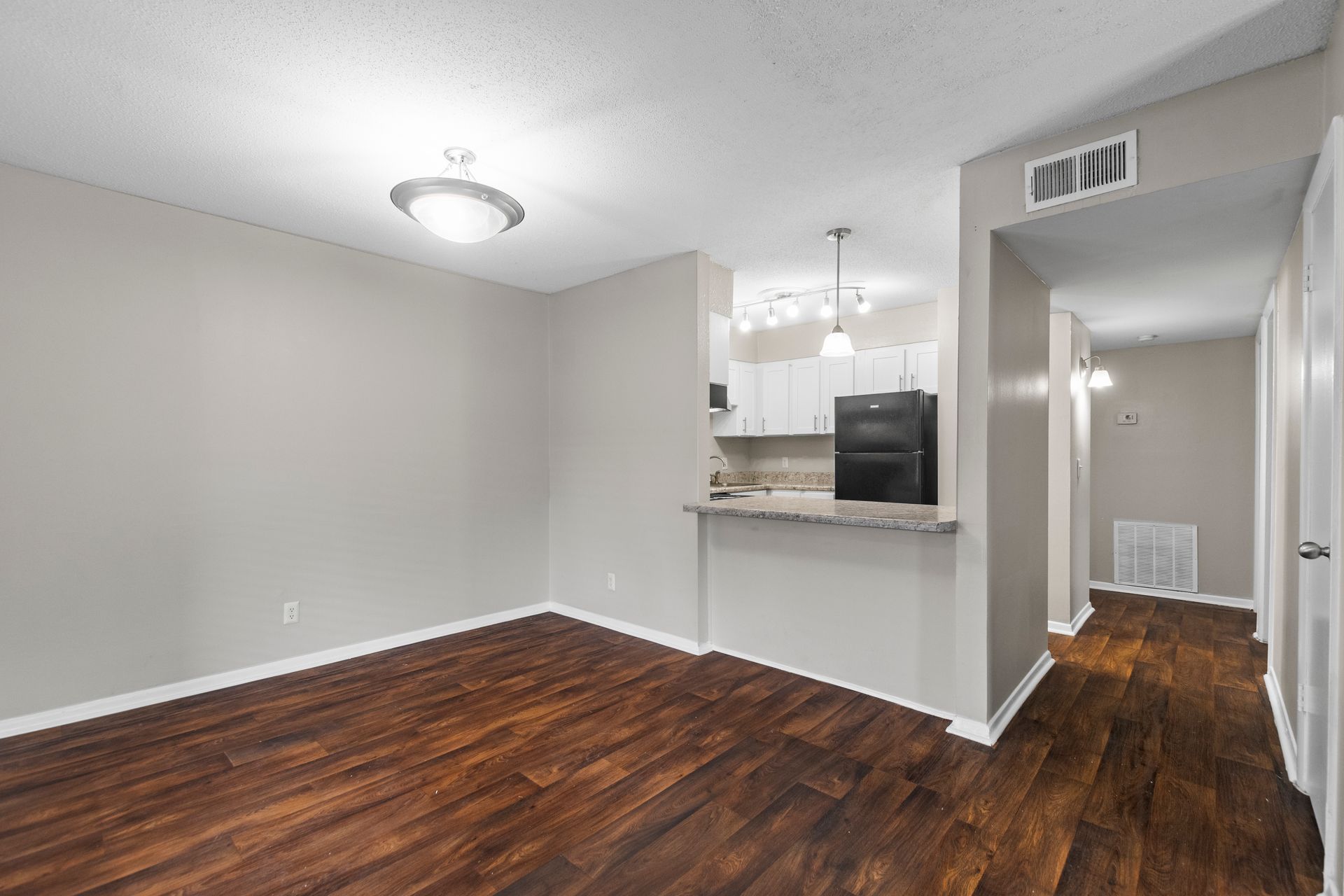 An empty living space featuring dark wood-look flooring, light grey walls, and a breakfast bar opening to the kitchen.