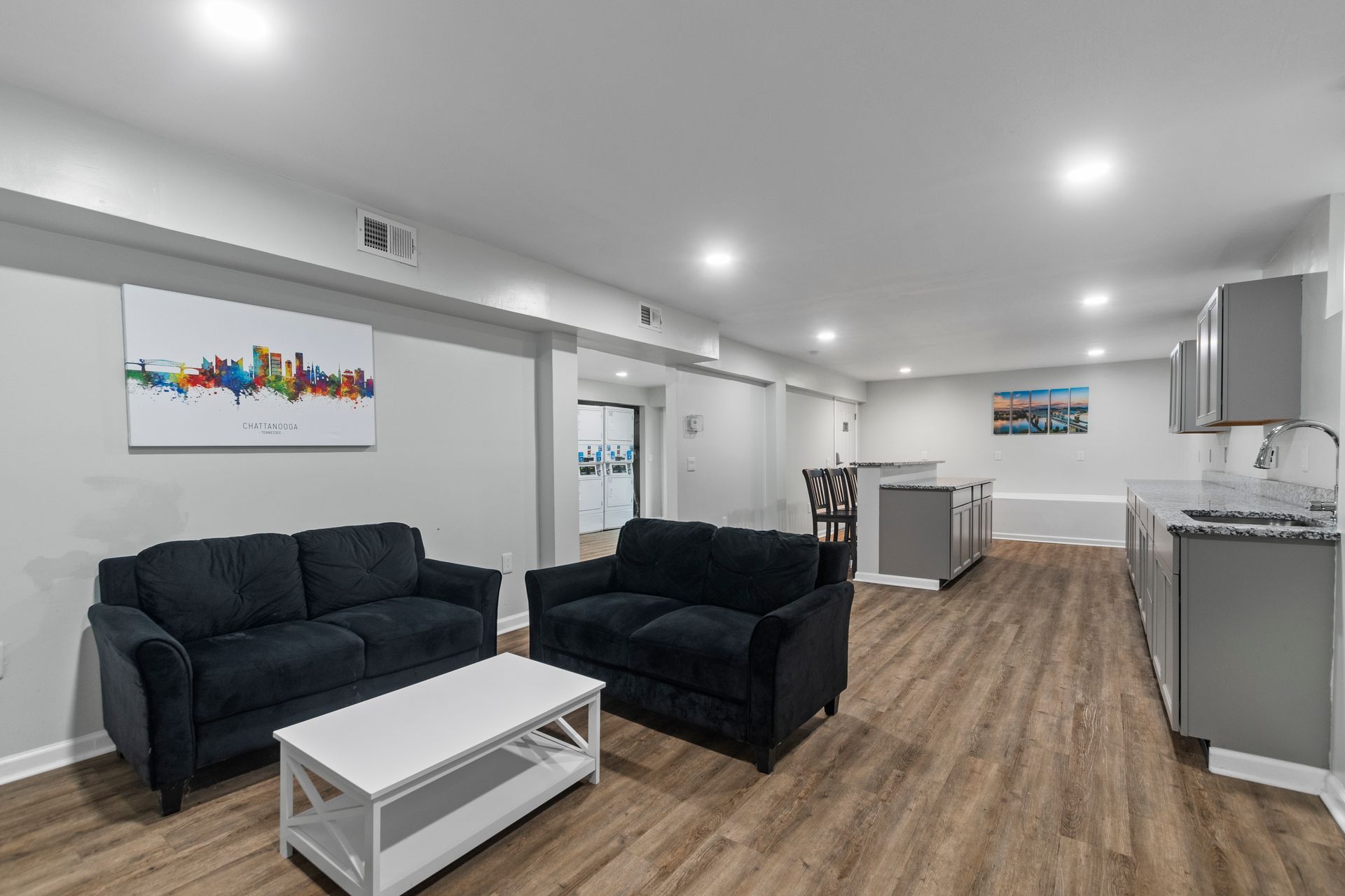 Modern living area with two dark sofas, a white coffee table, and an open kitchen with gray cabinets on wood floors.