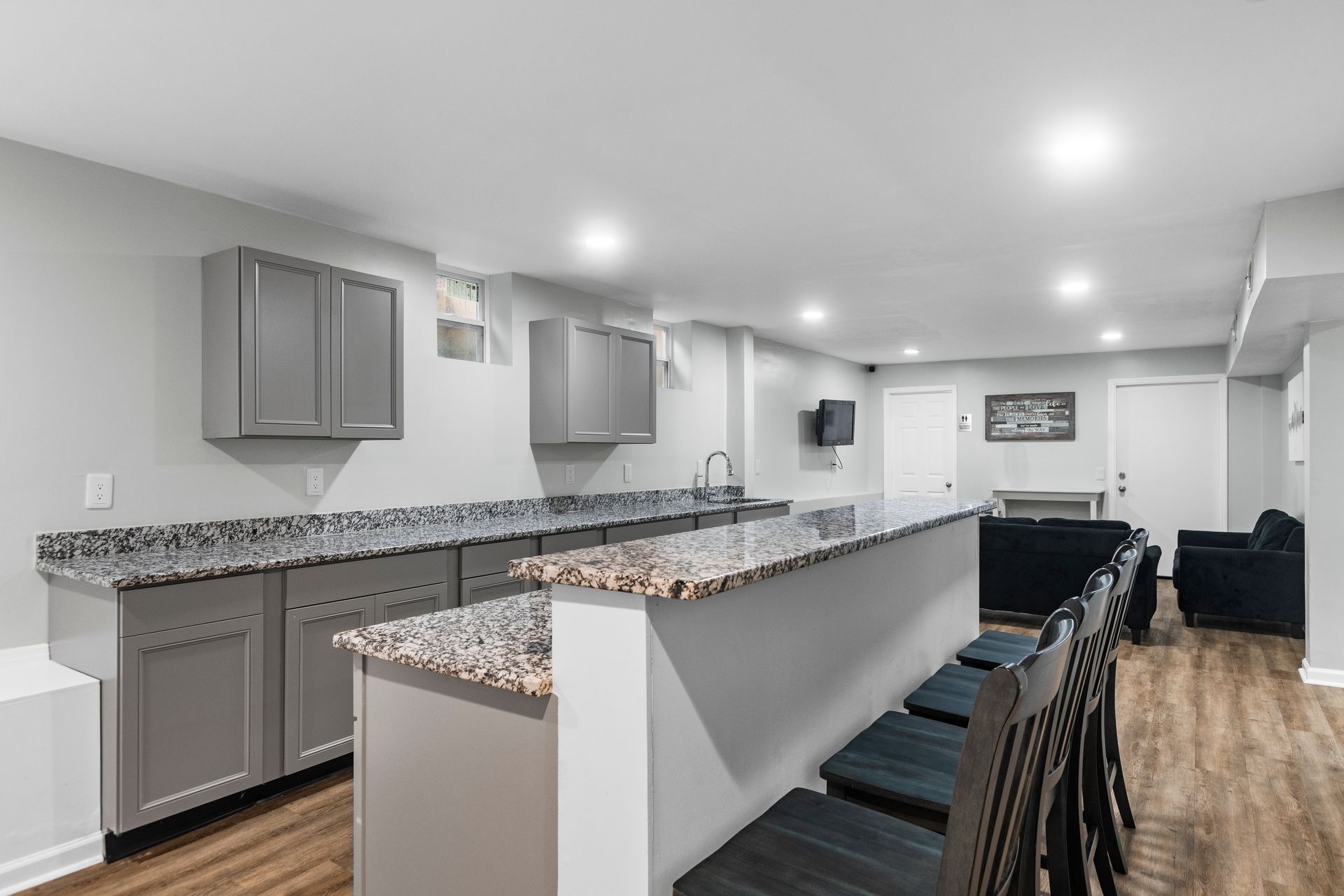 A finished basement with a kitchenettes area featuring grey cabinets, granite countertops, and bar seating for four.