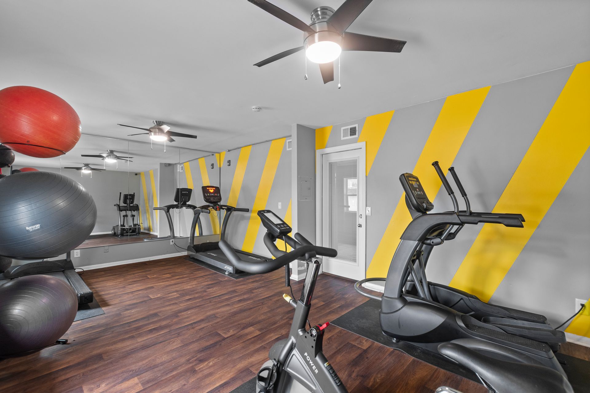 Small gym with an exercise bike, treadmill, yoga balls, and a yellow-and-gray striped wall.