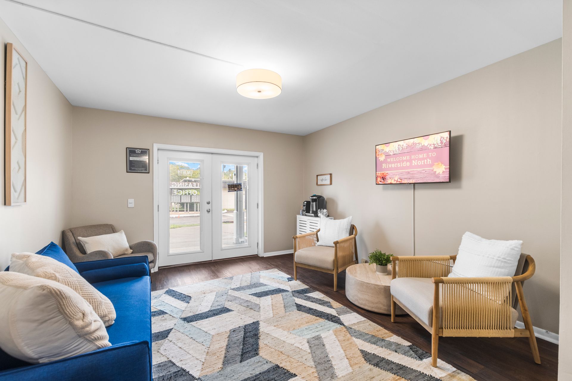 A modern waiting area with a blue sofa, light wood armchairs, a geometric rug, and a TV mounted on a beige wall.