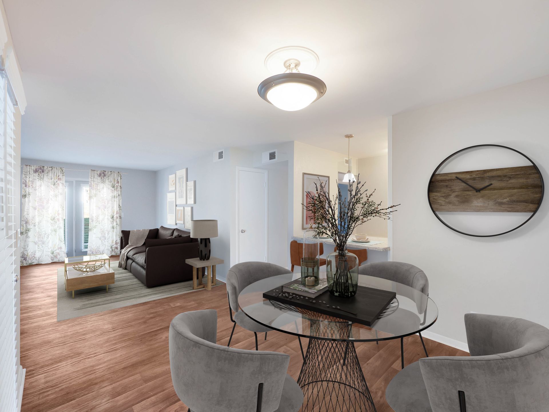 A dining area with a glass table and grey chairs opens into a living room with a brown sofa, light flooring, and wall clock.