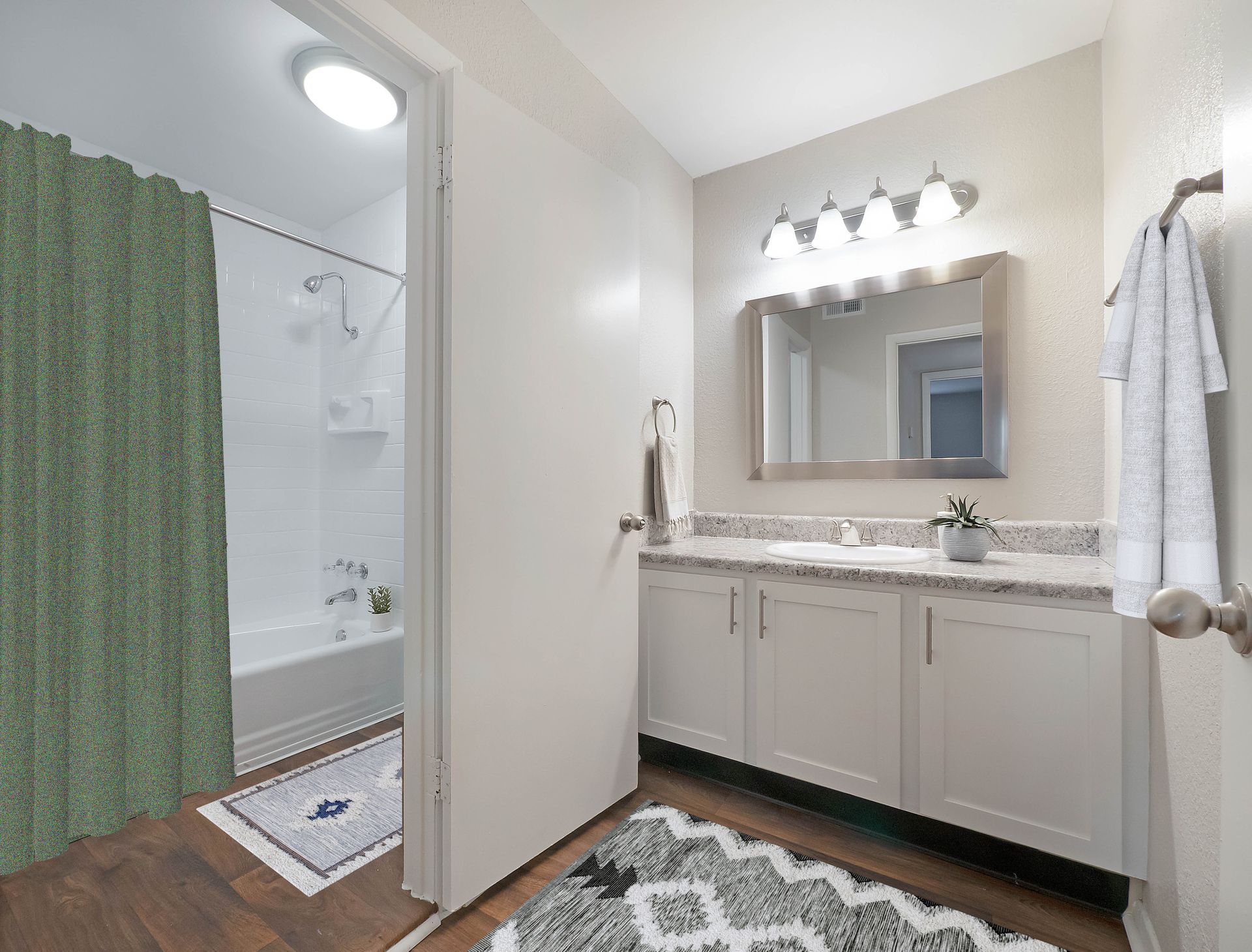 A modern bathroom featuring a white vanity with a mirror, a bathtub with a green curtain, and patterned area rugs.