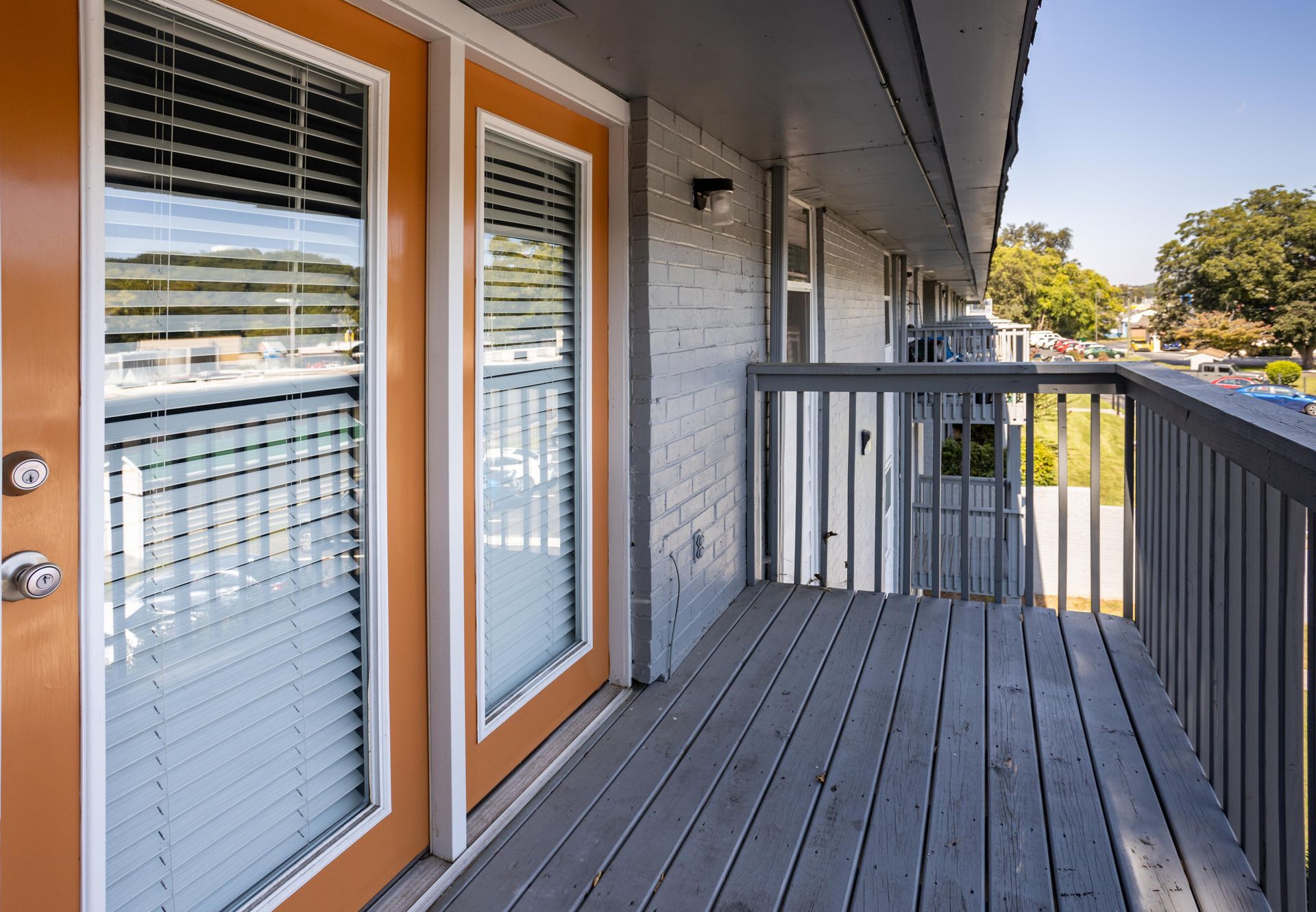 An apartment balcony with gray wooden floorboards and railing, beside two glass doors with orange frames.