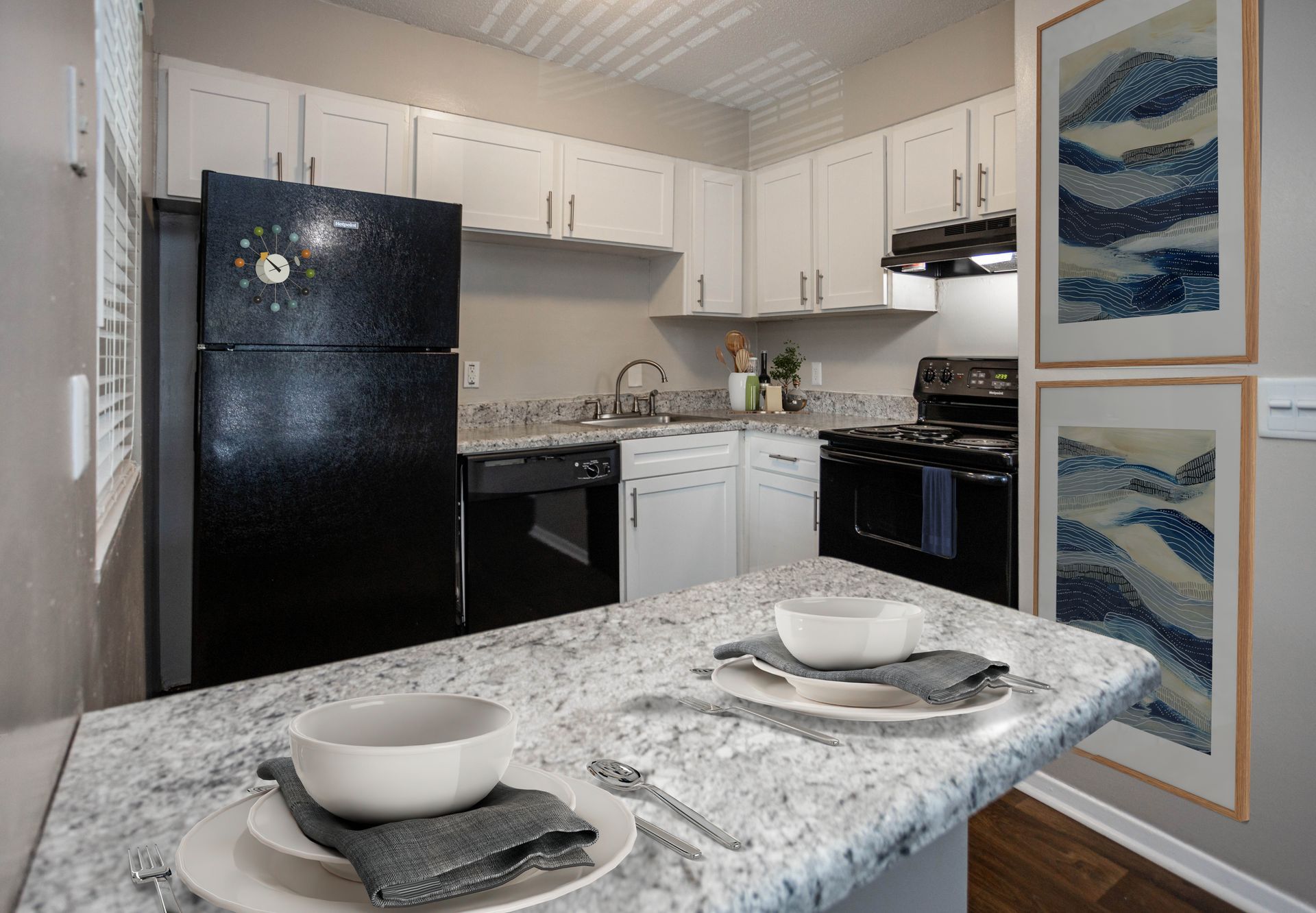 A modern kitchen featuring white cabinets, black appliances, speckled granite countertops, and two framed abstract prints.