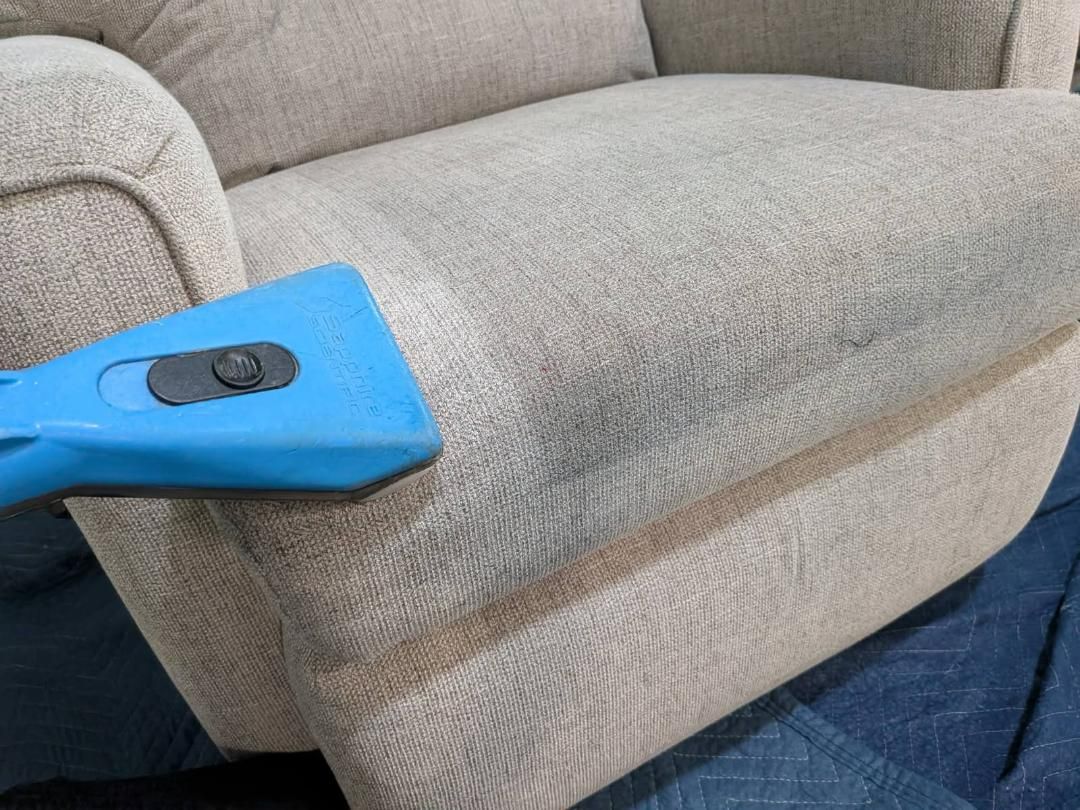 A person is cleaning a beige upholstered chair with a blue cleaning tool.