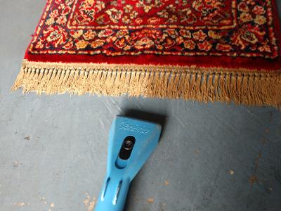 Red rug with fringe being cleaned by blue vacuum tool on a gray surface.