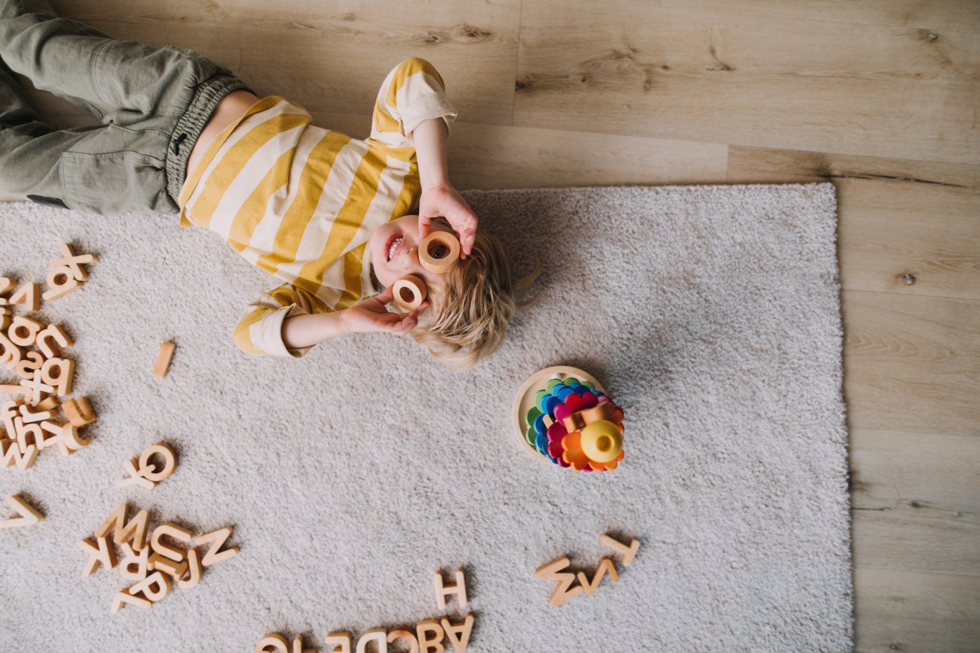 Boy lying on rug with wooden letters, holding donuts over eyes, smiling.