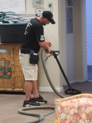 Man in a black shirt cleans a carpet with a vacuum cleaner. He is wearing shorts and a hat, indoors.