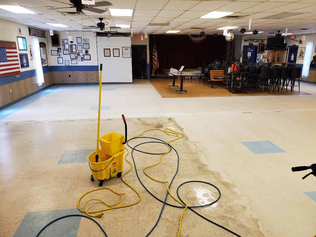 A room with wet floor being cleaned, yellow mop bucket and cord in the foreground. Stage, chairs, and flag visible.