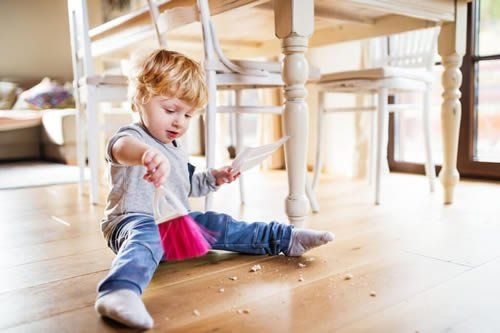 Blond toddler sweeping crumbs with a small pink broom under a white dining table.