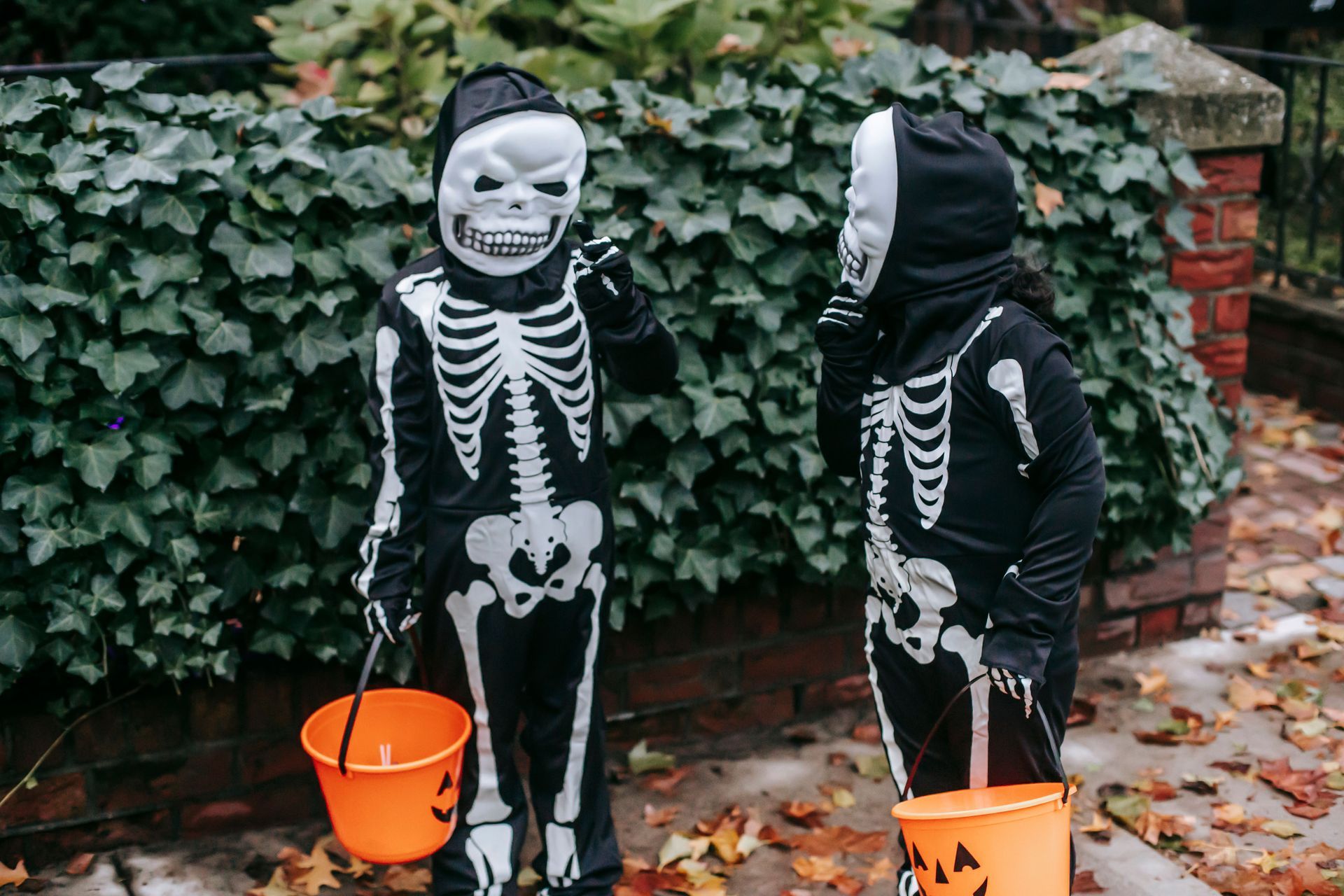 Kids dressed up as skeletons holding orange pumpkin buckets for candy collecting in front of a hedge