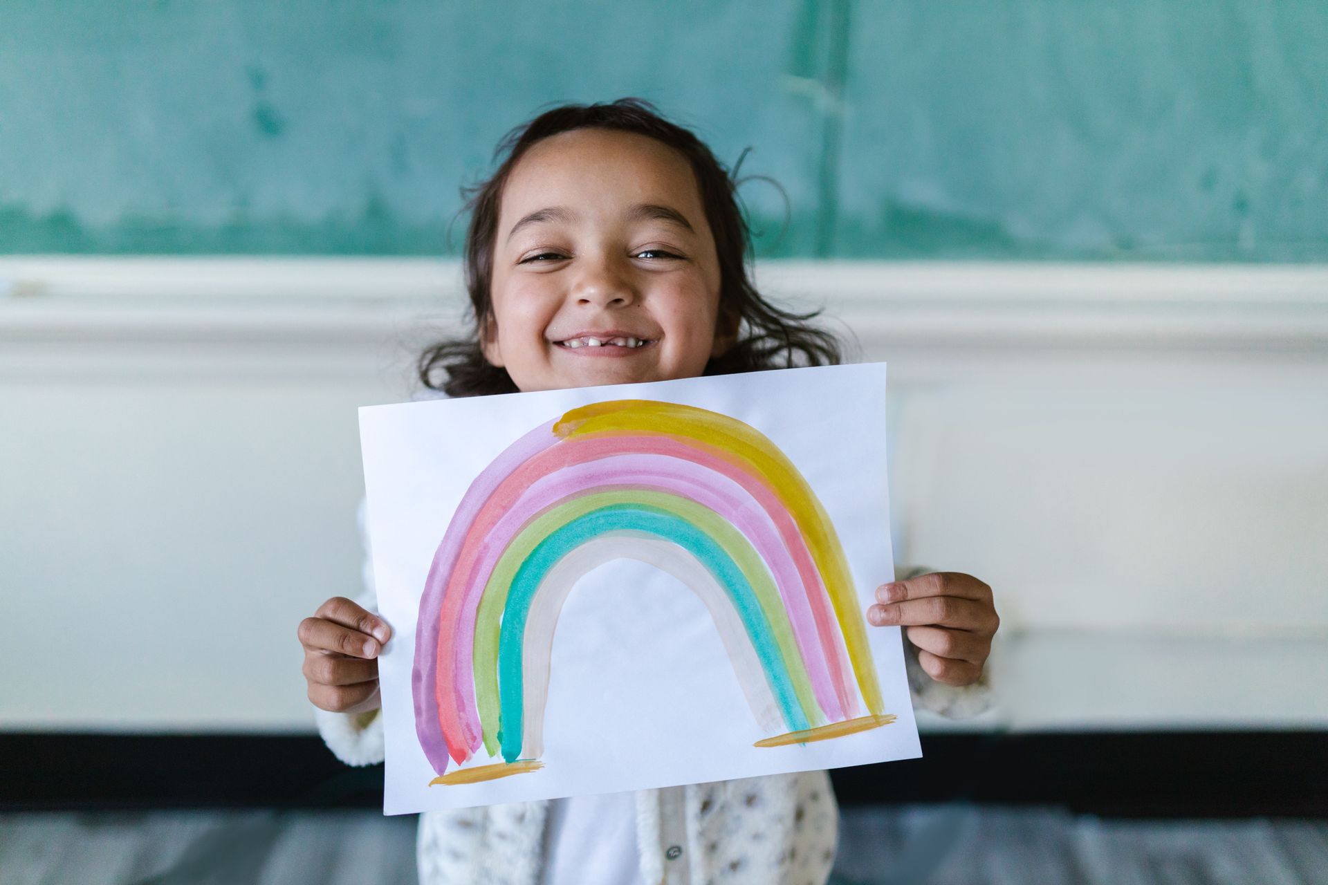 A young smiling girl holding a drawn picture of a rainbow.