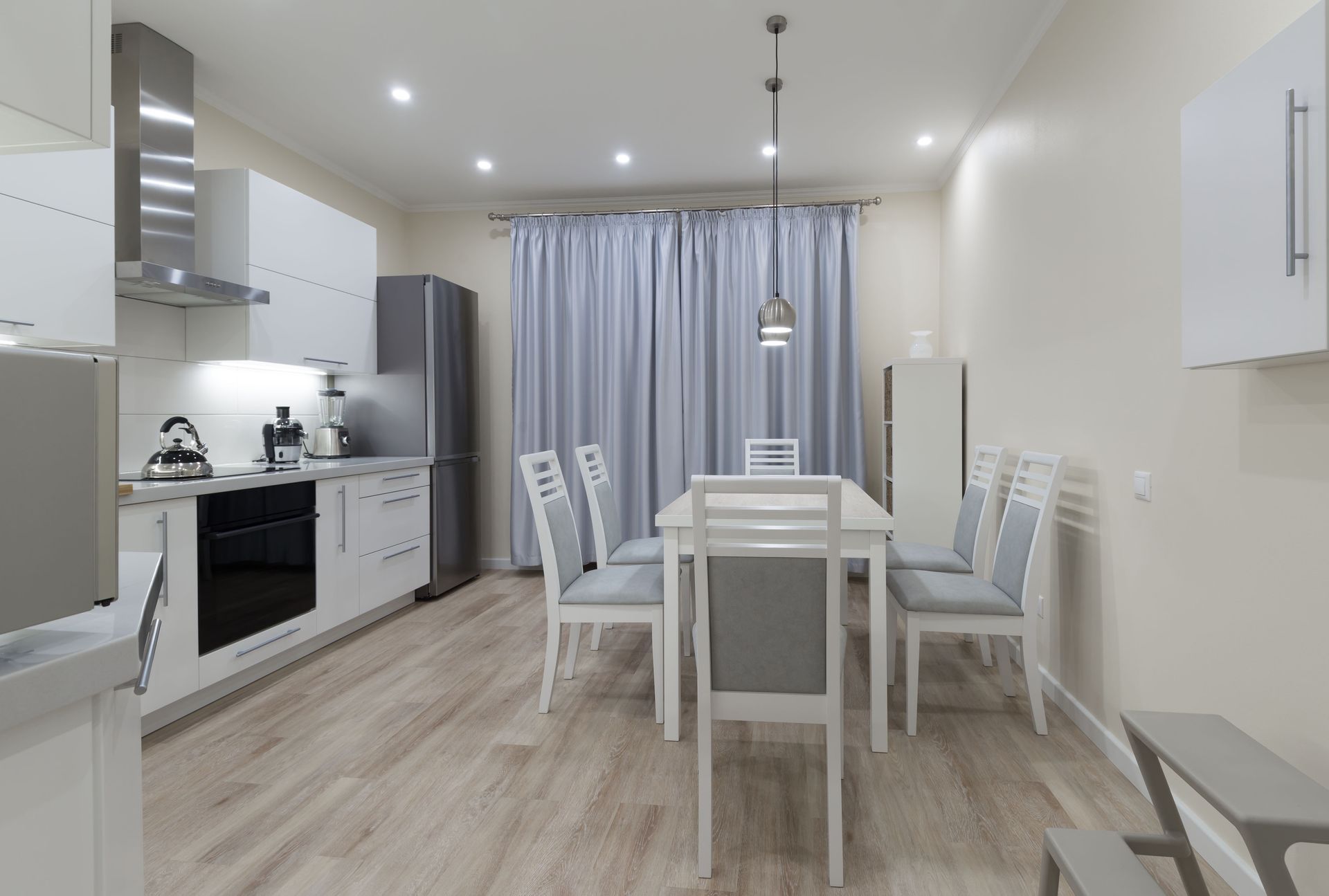 A modern, white-toned kitchen and dining area with a dining table, six chairs, stainless steel appliances, and wood floors.