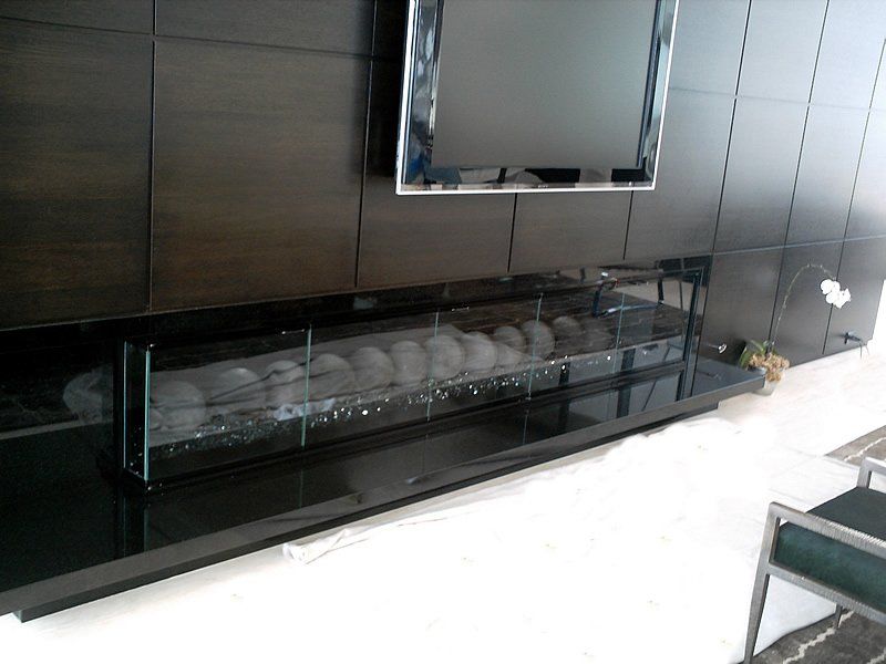 Modern fireplace with glass enclosure, black hearth, grey river rocks, beneath a mounted TV on a dark wooden wall.