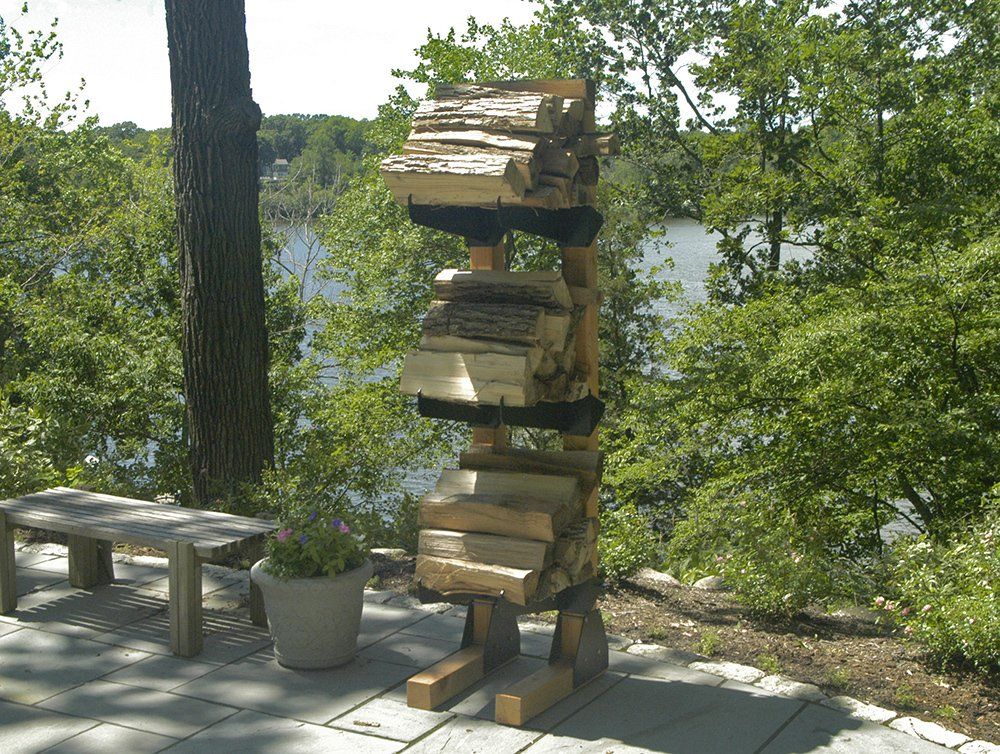 Sculpture of stacked firewood resembling a standing creature, next to a bench, overlooking a lake.