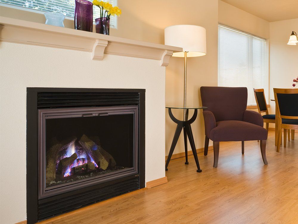 Fireplace in a cream-colored wall, with a mantle holding vases, and wood flooring, with a chair and table.
