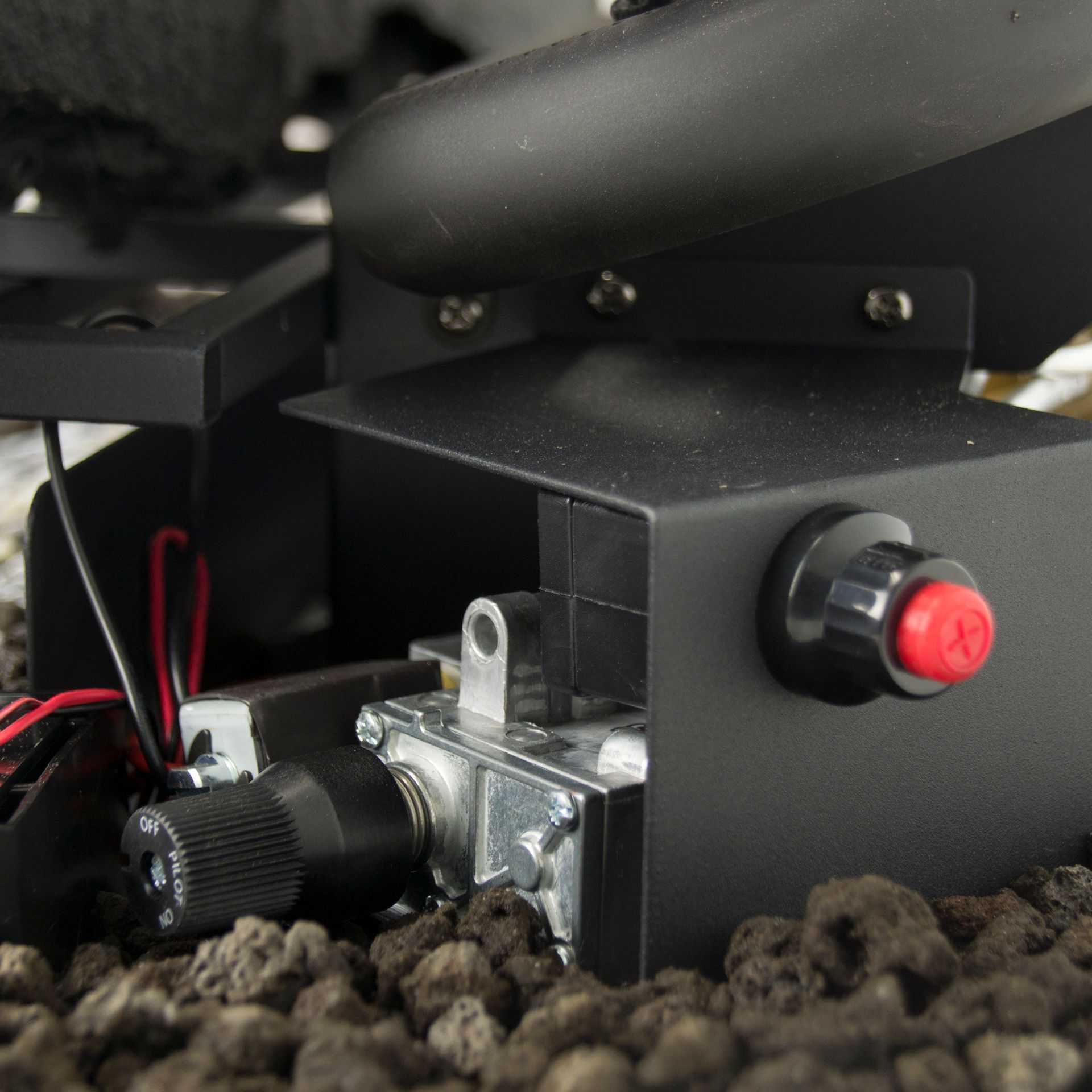 Black metal fire pit component with control knob, red button, and wiring, resting on lava rocks.