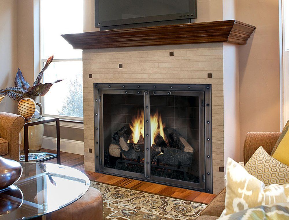 Fireplace with flames, metal doors, wood mantel, beige tile, rug, and furniture in a living room.