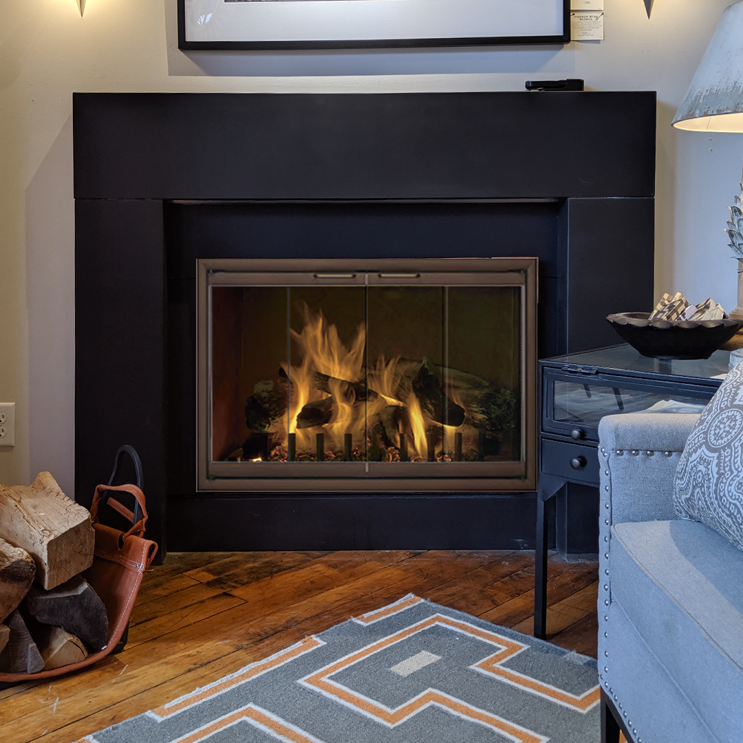 Fireplace with flames behind glass doors, black frame. Stacked firewood basket, rug, and furniture in room.