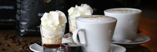 Twee witte kopjes en een klein glas, alle met slagroom erop, staan ​​op een tafel met koffiebonen en een zak koffie.