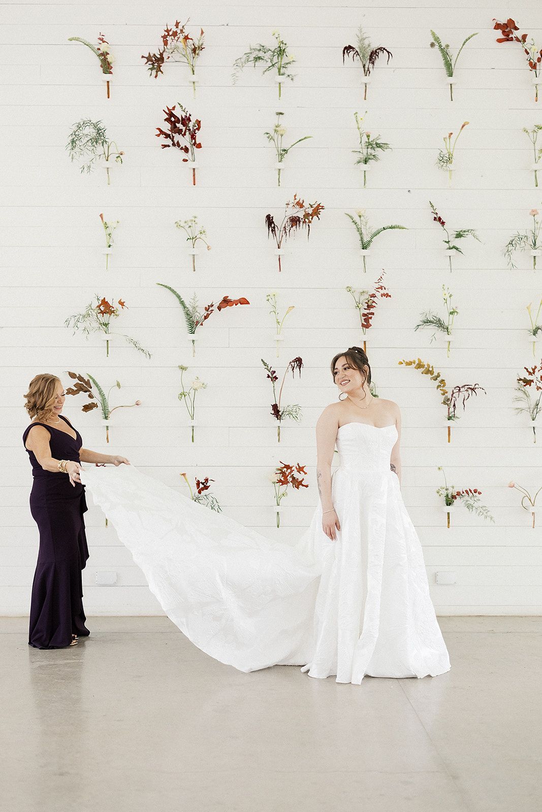 Bride in white gown with attendant holding long veil in a minimalist floral wall setting