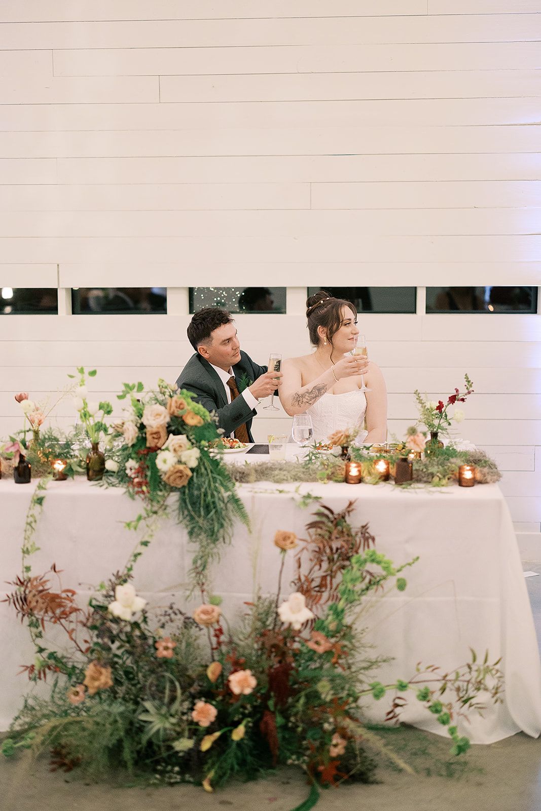 Wedding couple seated at a floral-decorated table, smiling with champagne glasses in a bright reception hall