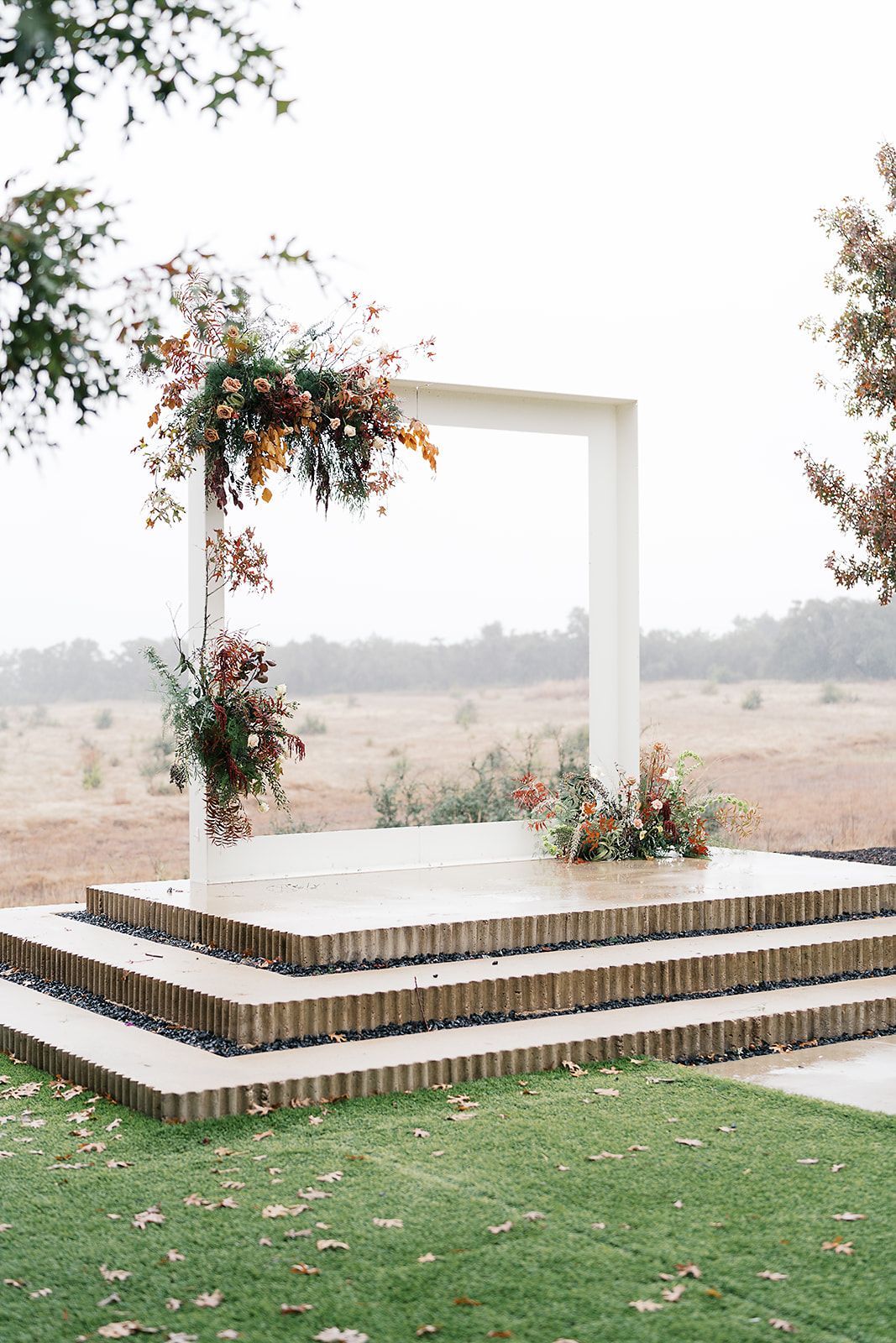 Outdoor white wedding arch with floral vines on a stepped platform in a grassy field
