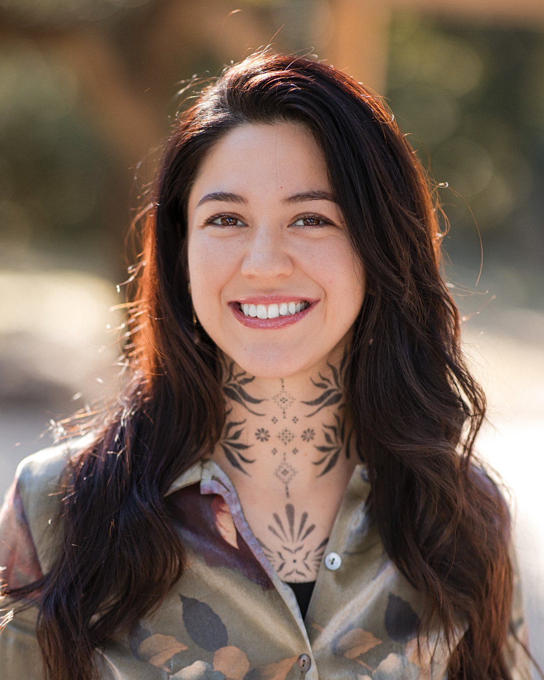 Smiling person with dark hair wearing a patterned shirt outdoors in soft sunlight