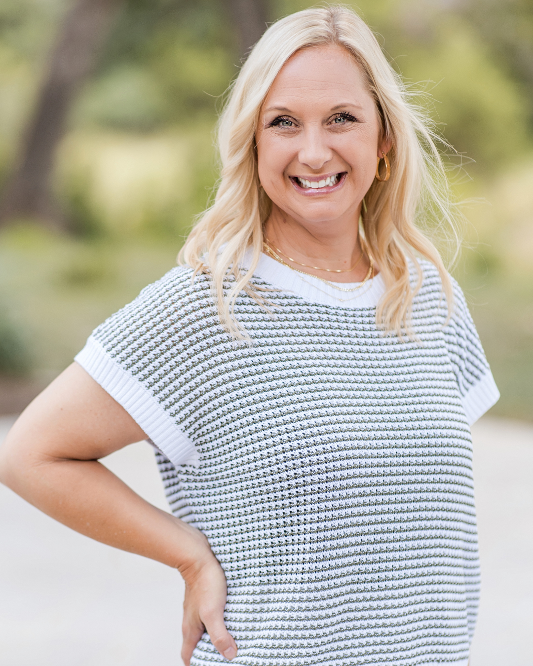Smiling blonde woman in a gray patterned top standing outdoors with one hand on her hip