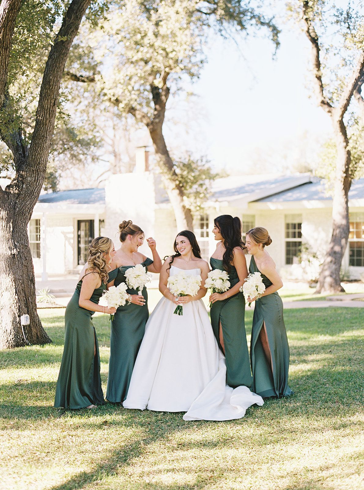 Bride with four bridesmaids in green dresses standing outdoors in front of a house, holding bouquets.