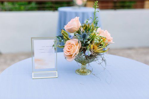 Floral centerpiece with pale pink roses and greenery on a round table beside a framed sign