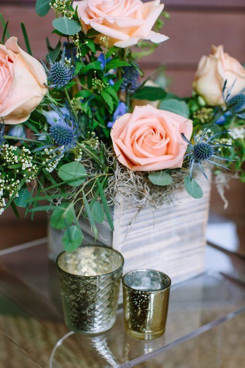 Peach roses and greenery in a white vase with two gold candle holders on a glass table.