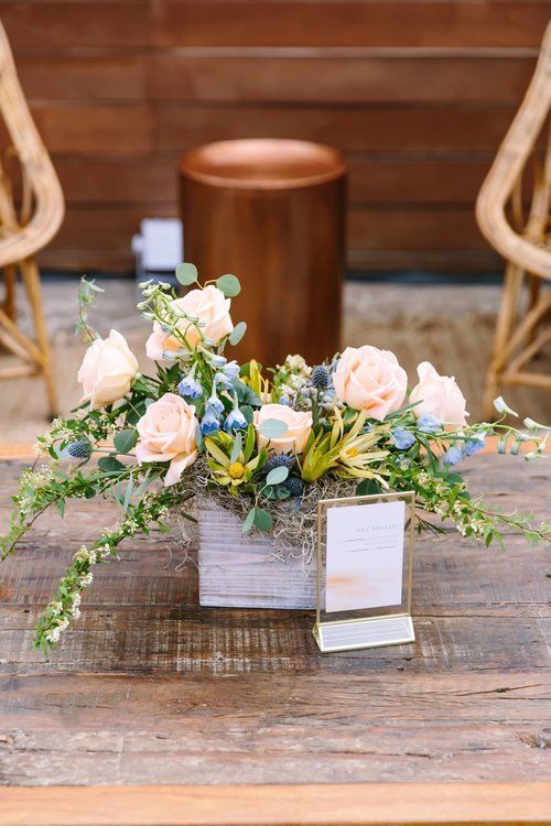 Floral arrangement with pale pink roses and greenery in a wooden box on a rustic table, with a name card behind it