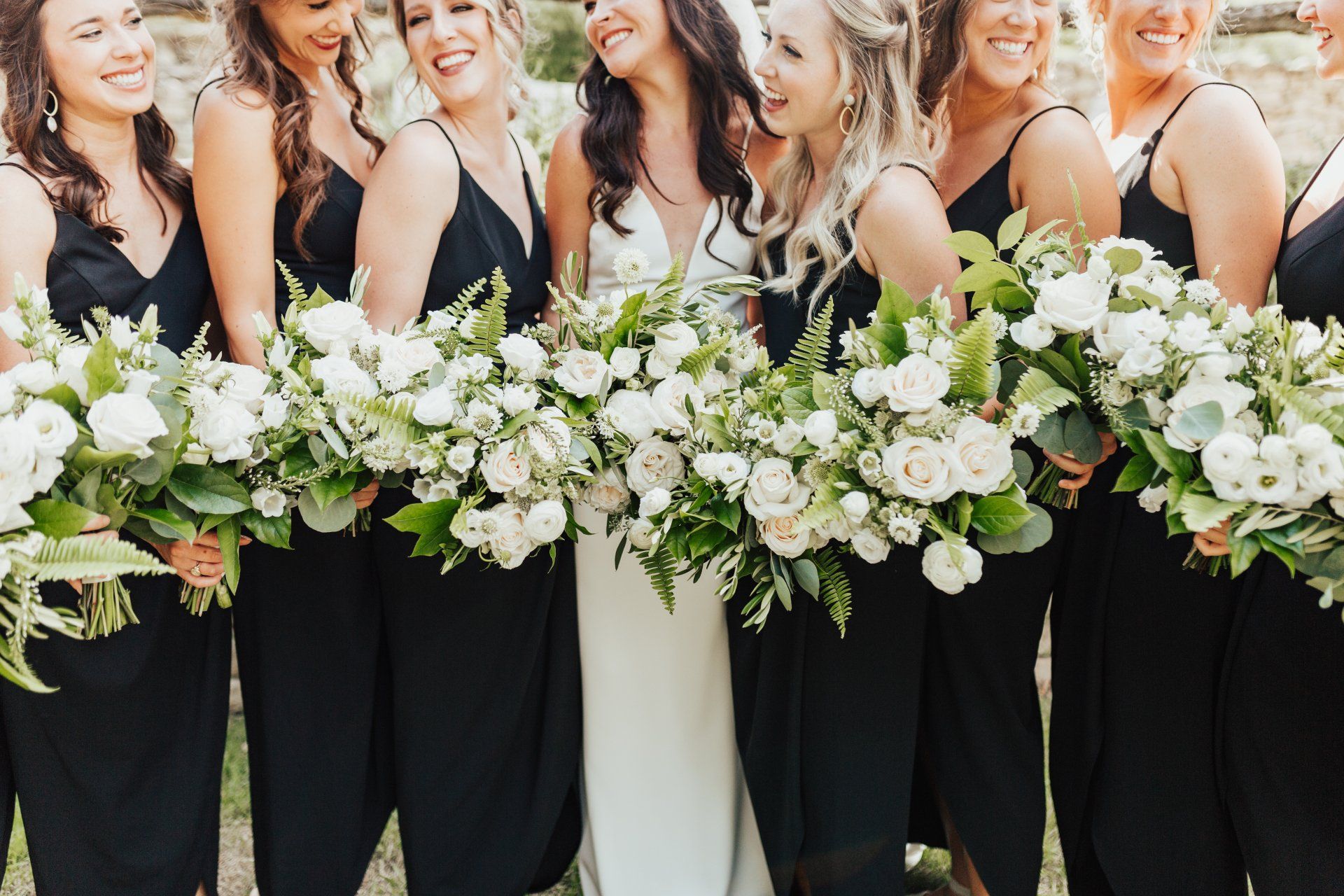 Bridesmaids in black dresses and one in white hold white floral bouquets at an outdoor wedding.