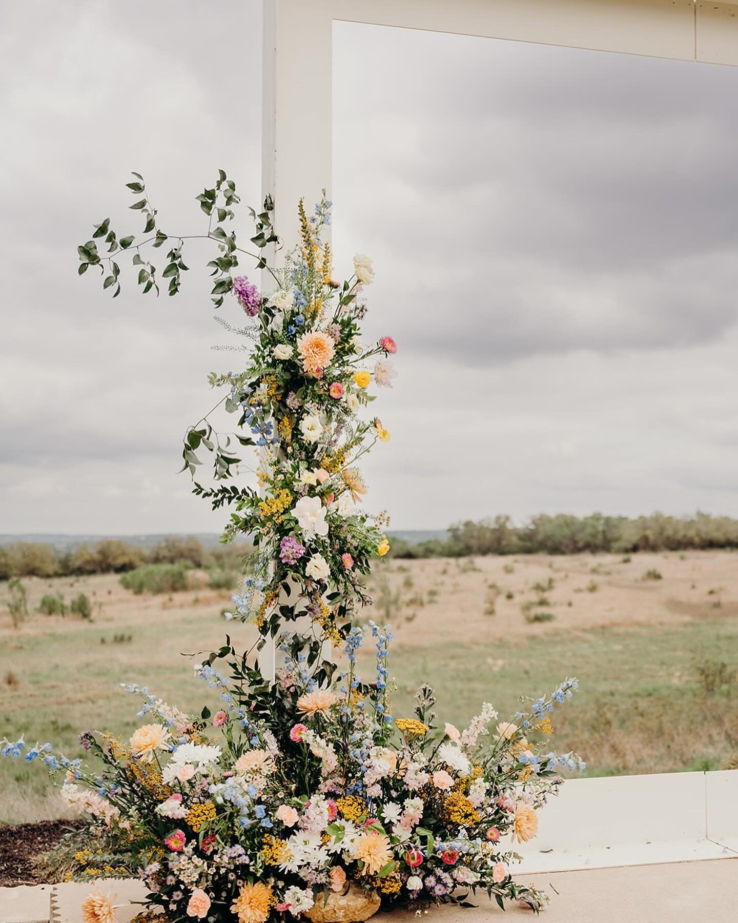 Tall floral arrangement with pastel flowers and greenery in an outdoor field under a cloudy sky