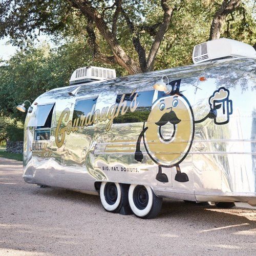 Silver food trailer with a cartoon logo parked under trees on a roadside, with dual wheels and rooftop vents