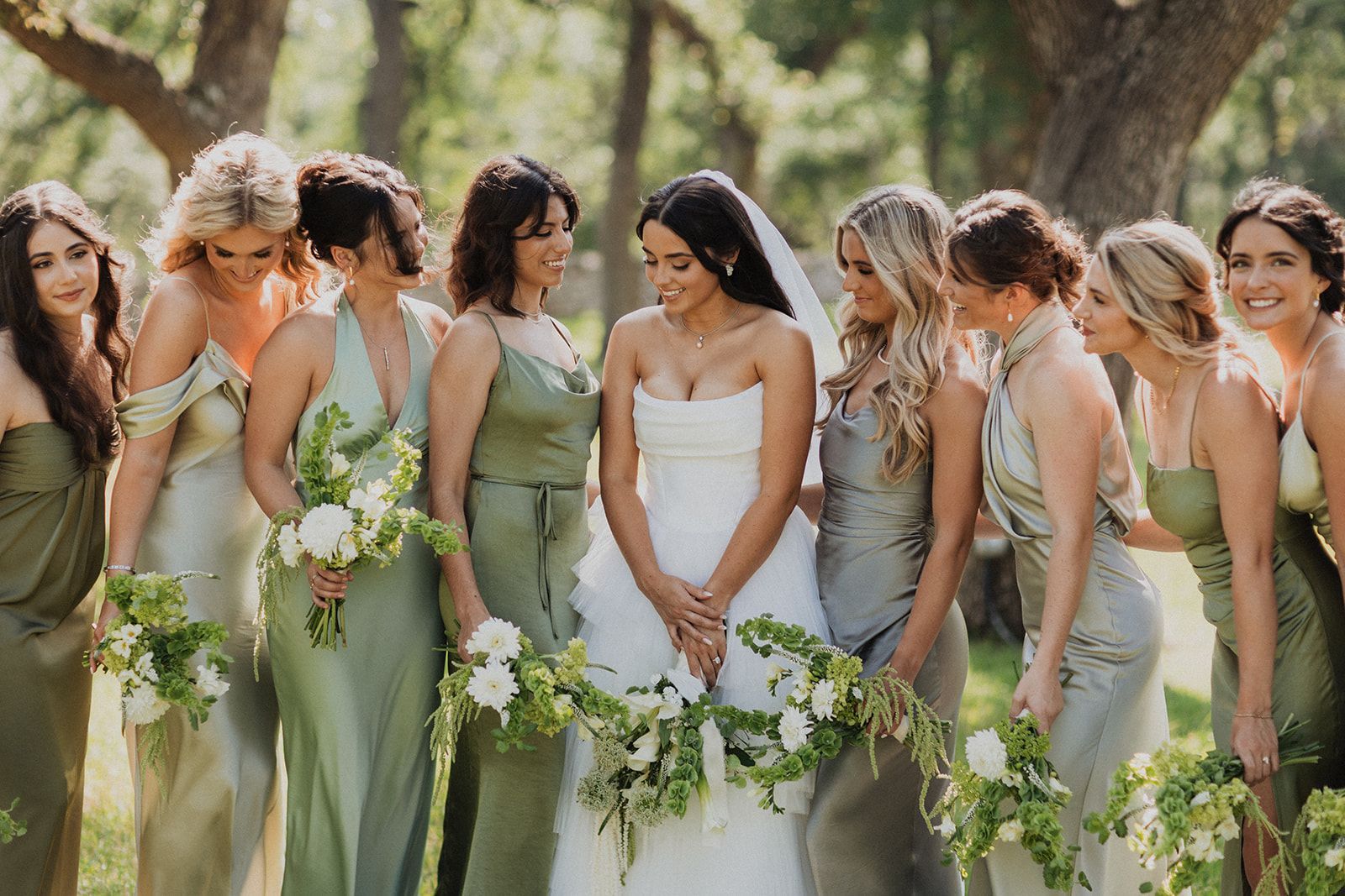 Bride in white dress surrounded by bridesmaids in sage green dresses holding bouquets outdoors