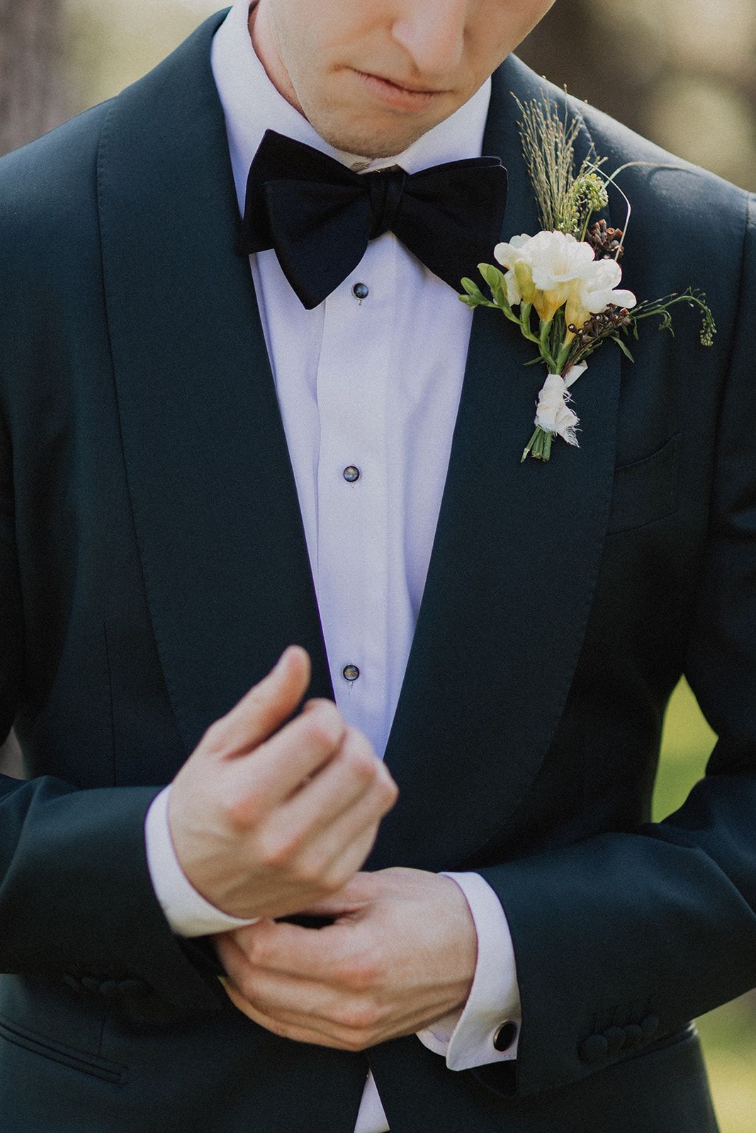 Man in a black tuxedo adjusting his cuff, wearing a white boutonniere on the lapel.