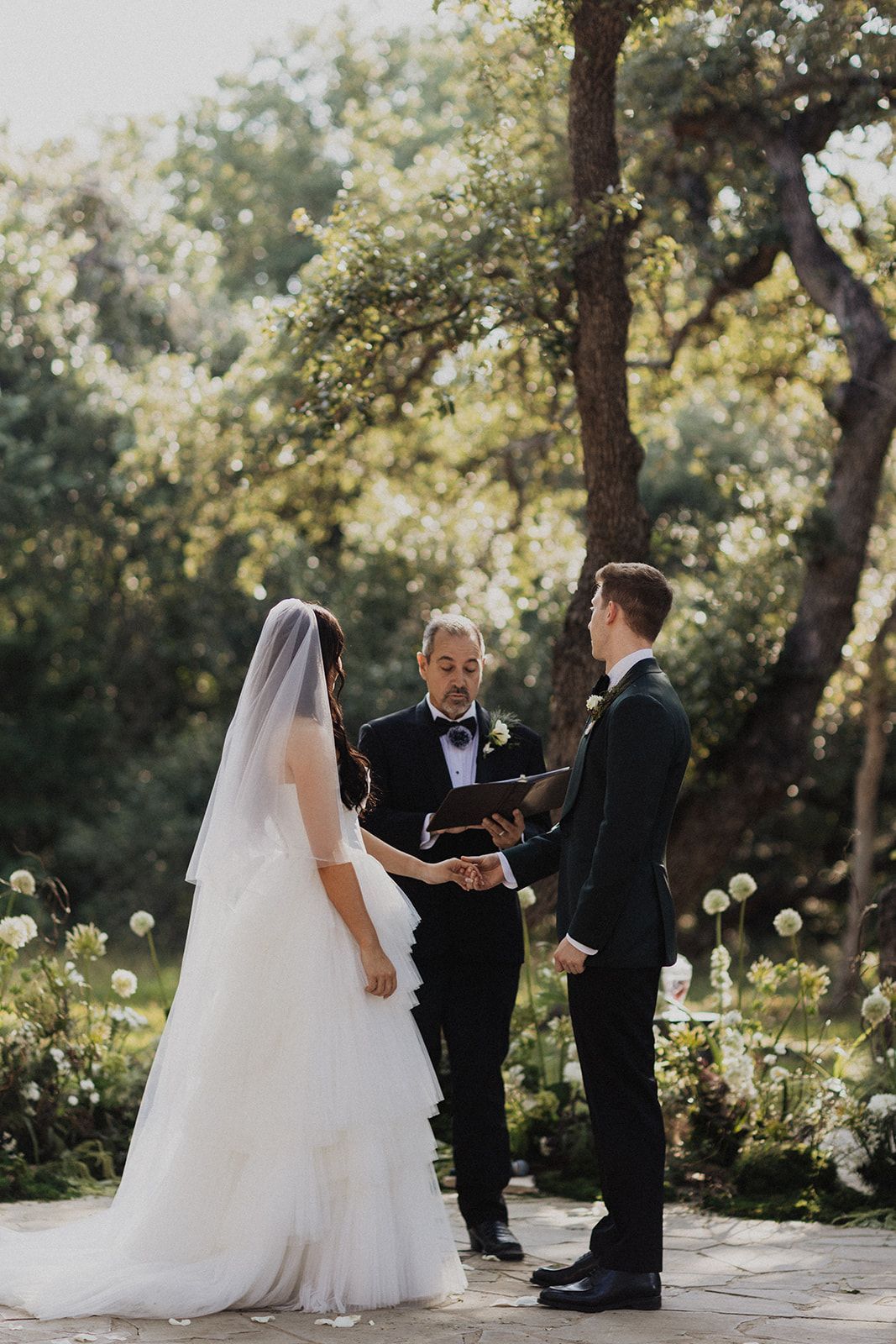 Bride and groom stand with an officiant during an outdoor wedding ceremony in a garden.