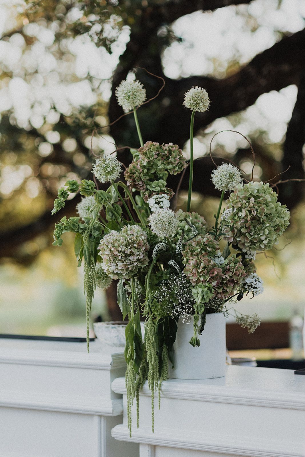 White vase of green and white flowers on a white ledge, with a blurred outdoor background