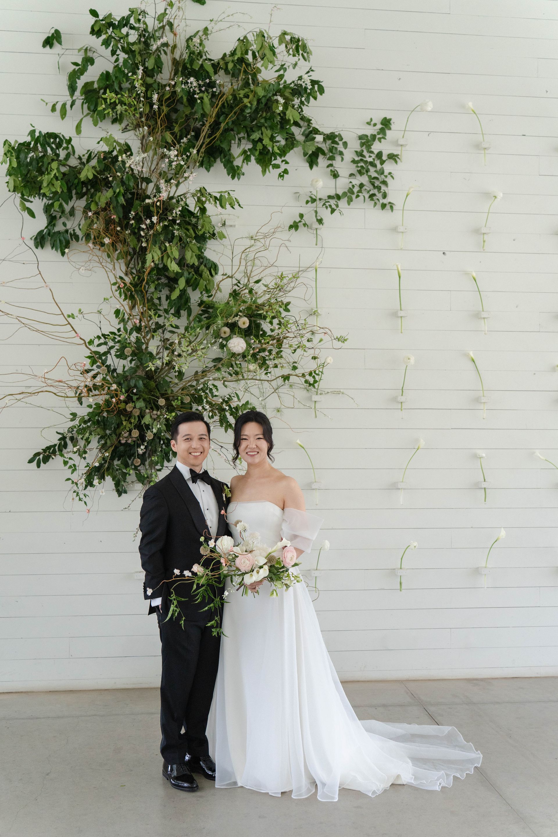 Wedding couple posing by a white wall with green vines, bride in white gown and groom in black suit