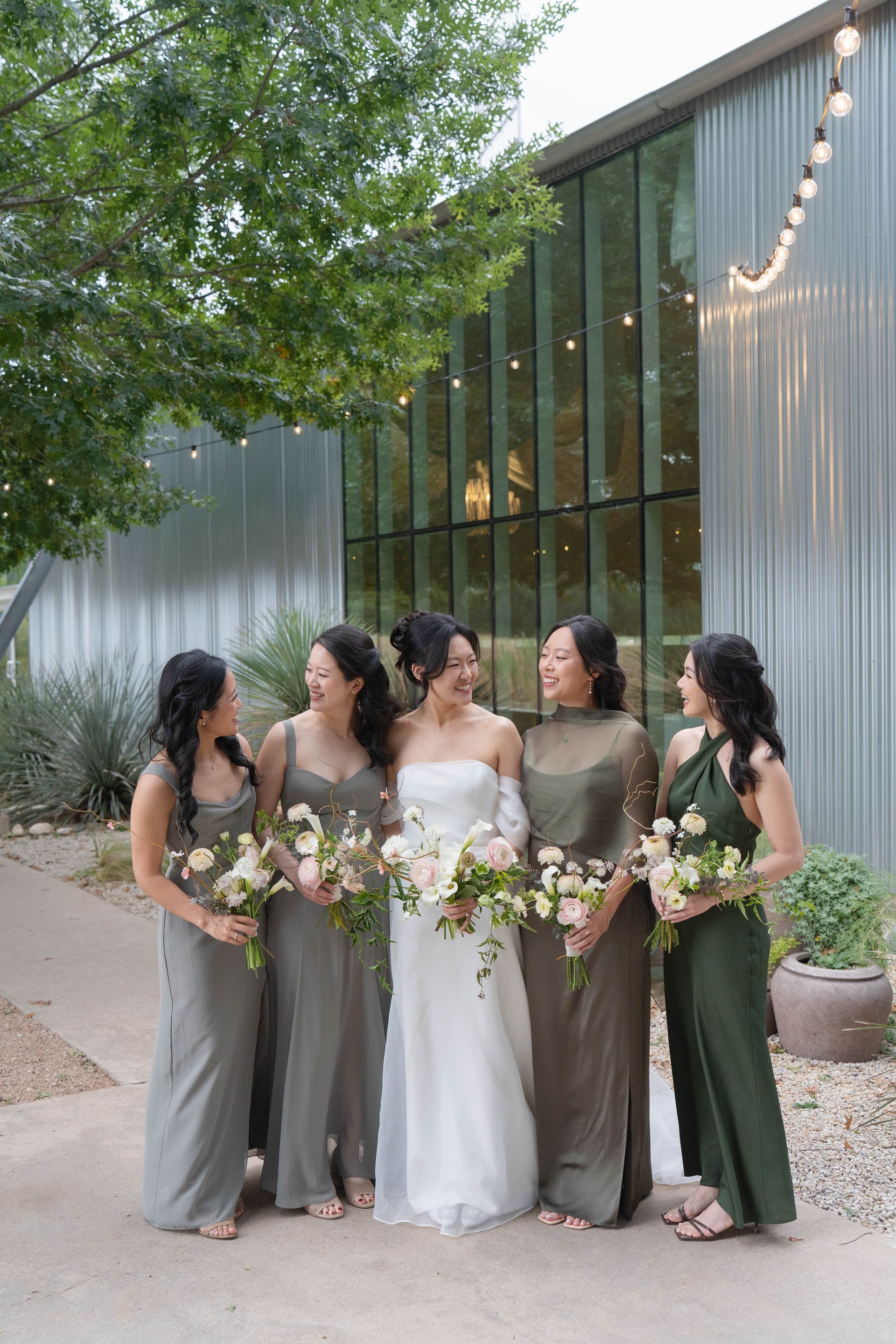 Five women in formal dresses holding bouquets outside a modern building at dusk