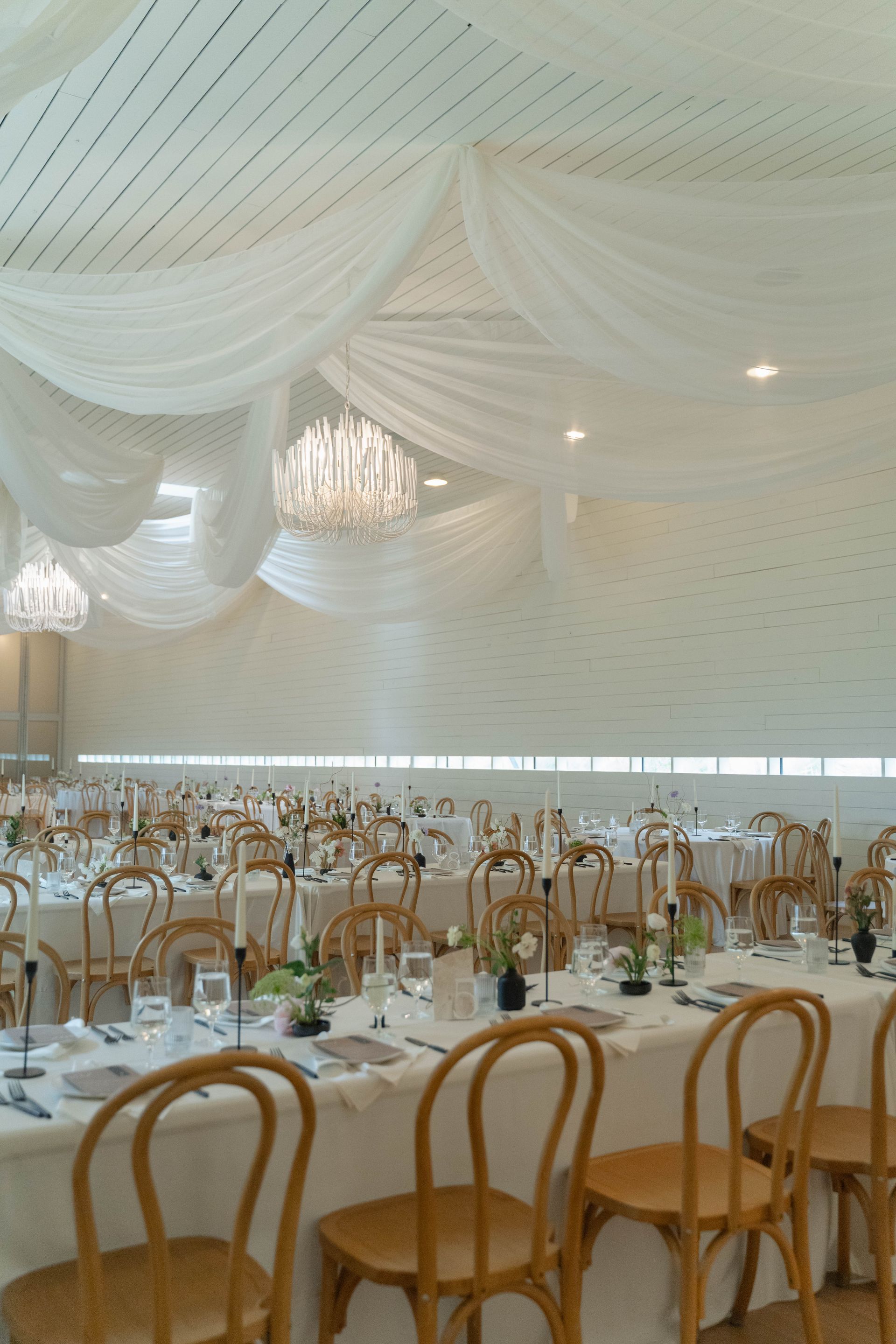 Elegant banquet hall with white draped ceiling, chandeliers, and rows of wooden chairs and tables set for an event