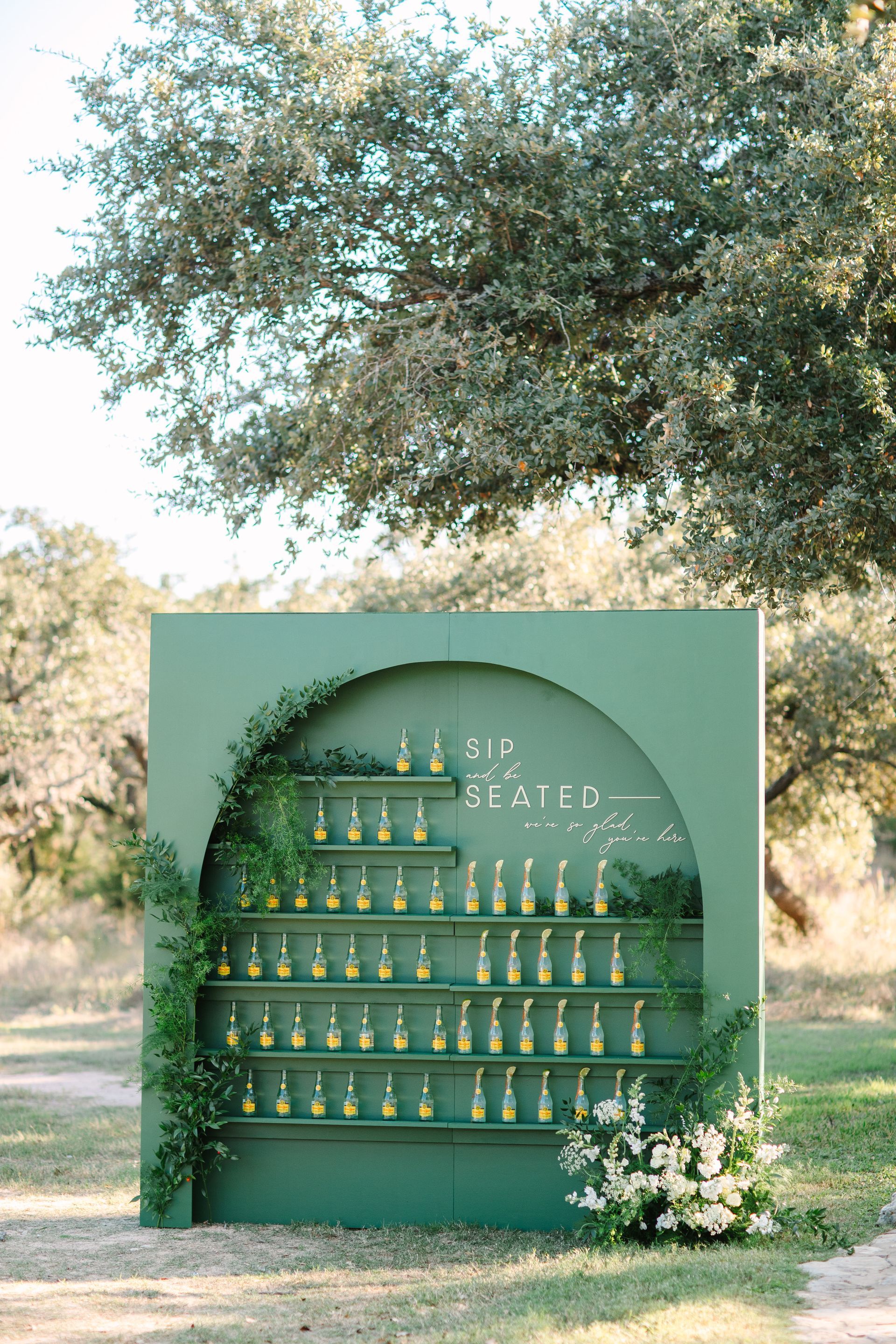 Green outdoor display of shelves with many small bottles and flowers beneath a tree