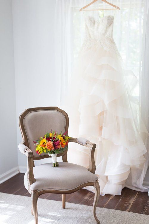 Wedding dress hanging beside a vintage chair with a colorful bouquet in a bright room